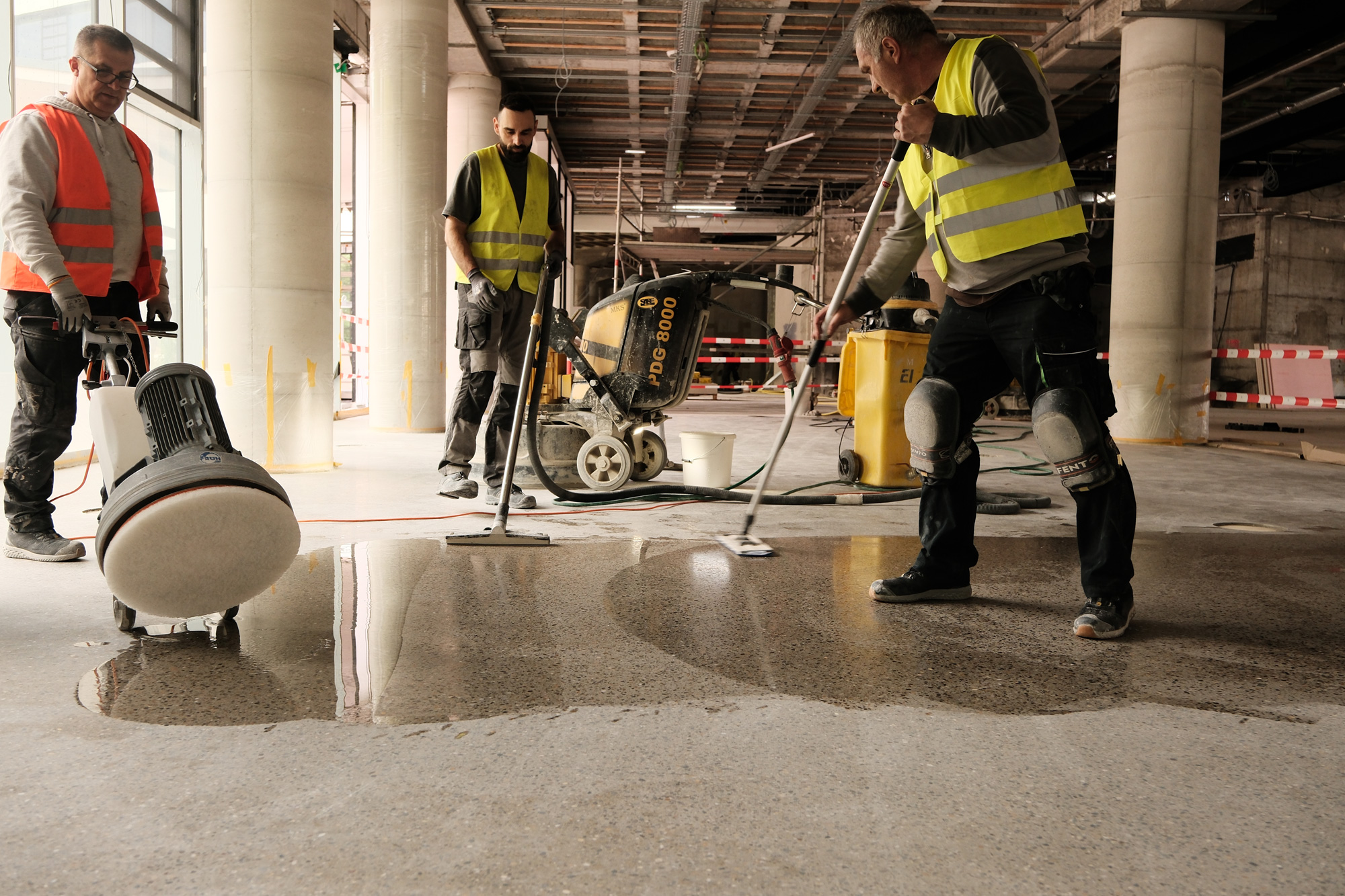 Drei Arbeiter beim Schleifen eines Sichtestrichbodens in Terrazzo-Optik, die mit Schleifmaschinen die Oberfl&auml;che bearbeiten und gl&auml;tten.